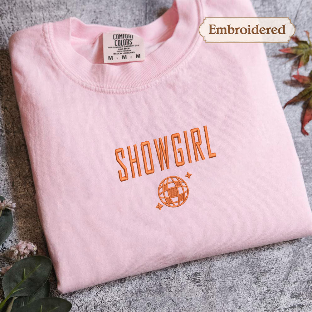 A pink t-shirt with the text "SHOWGIRL" and a globe embroidered on it, along with a label indicating it is embroidered.