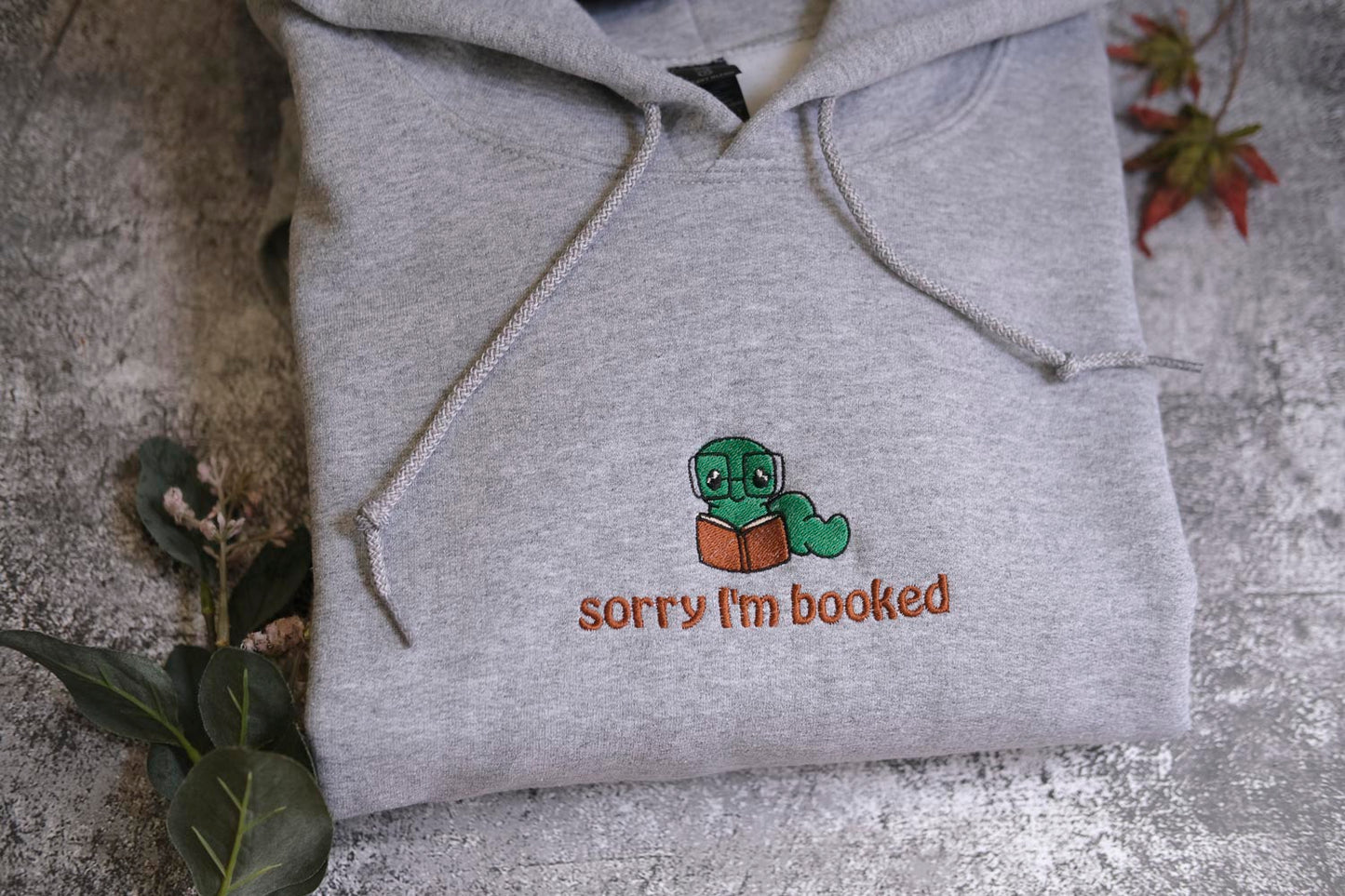 A gray hoodie with a small cartoon character and text printed on it, placed on a surface with some leaves and a plant.
