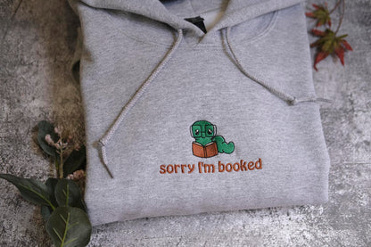 A gray hoodie with a small cartoon character and text printed on it, placed on a surface with some leaves and a plant.