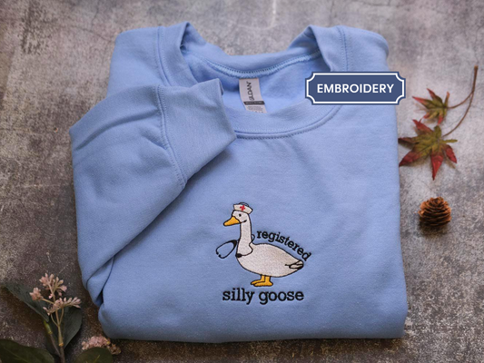 a light blue sweatshirt with a cartoon duck wearing a nurse's hat and the text "registered silly goose" printed on it. The sweatshirt is laid out on a gray surface, with some leaves and pine cones scattered around it.