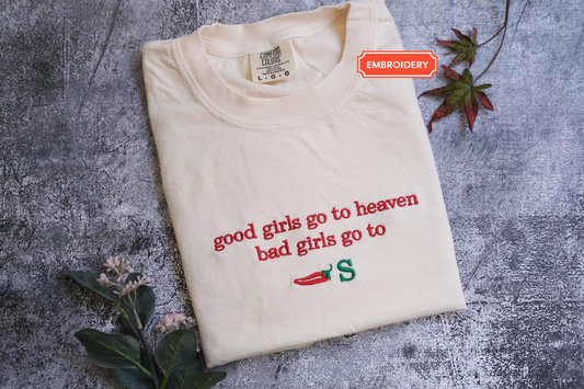 a white t-shirt with a red and green embroidered phrase that reads "good girls go to heaven bad girls go to S". The t-shirt is placed on a gray surface, and there are some leaves and a red flower nearby.