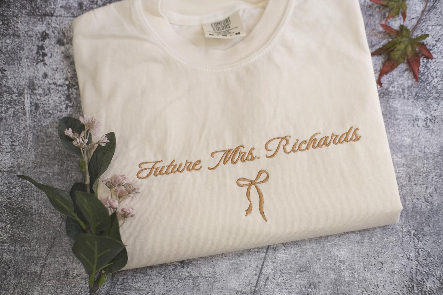 A white t-shirt with the text "Future Mrs. Richards" embroidered on it, along with a small gold ribbon bow. The t-shirt is placed on a gray surface, and there are some small pink flowers and a leaf nearby.