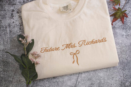 A white t-shirt with the text "Future Mrs. Richards" embroidered on it, along with a small gold ribbon bow. The t-shirt is placed on a gray surface, and there are some small pink flowers and a leaf nearby.