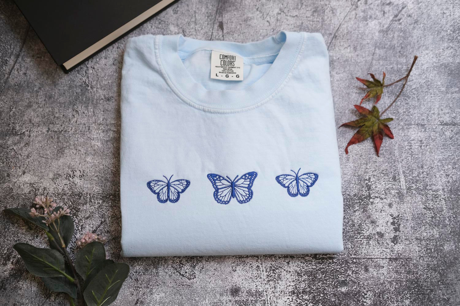 a light blue t-shirt with three blue butterflies printed on it, placed on a gray surface alongside a book and some leaves.