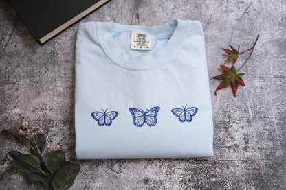 a light blue t-shirt with three blue butterflies printed on it, placed on a gray surface alongside a book and some leaves.