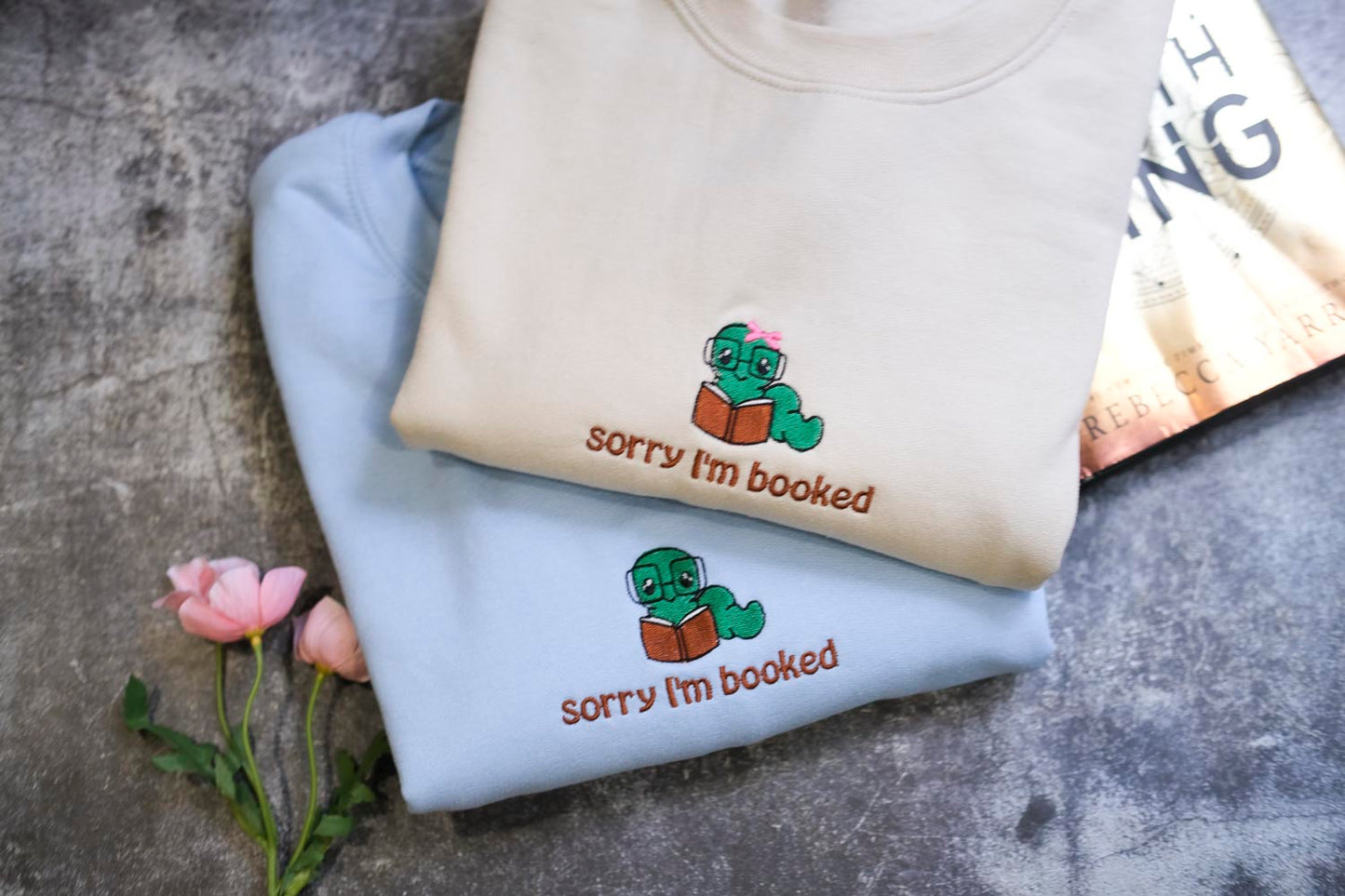 two light blue sweatshirts with a cartoon image of a green frog wearing glasses and holding a book, along with the text "Sorry I'm booked" printed on them.