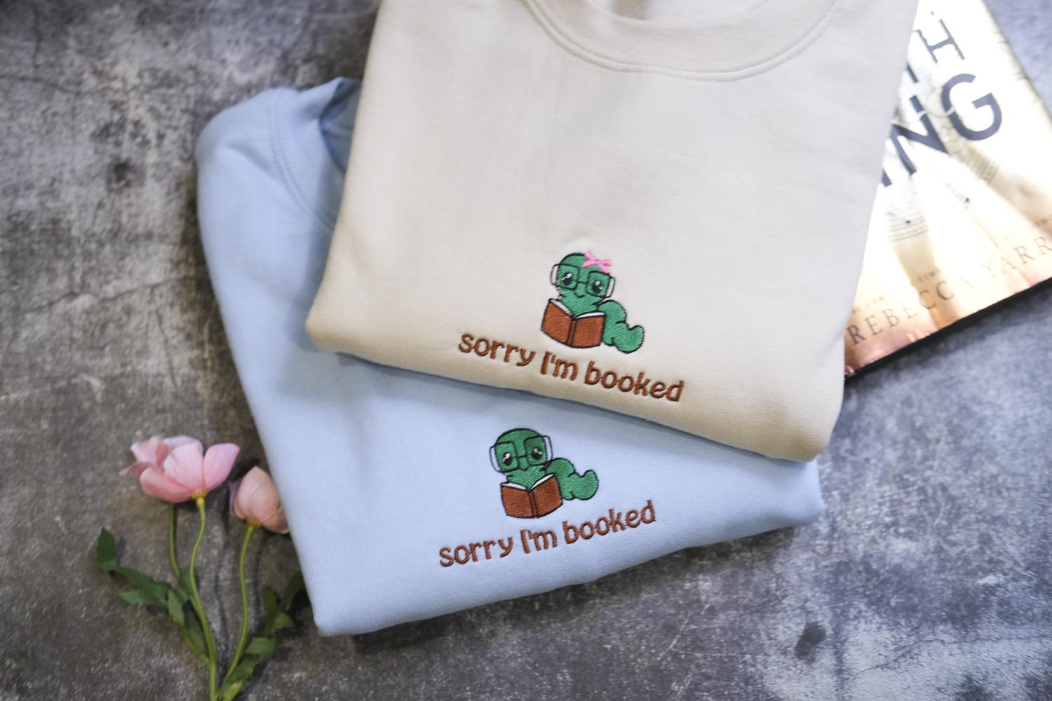 two light blue sweatshirts with a cartoon image of a green frog wearing glasses and holding a book, along with the text "Sorry I'm booked" printed on them.