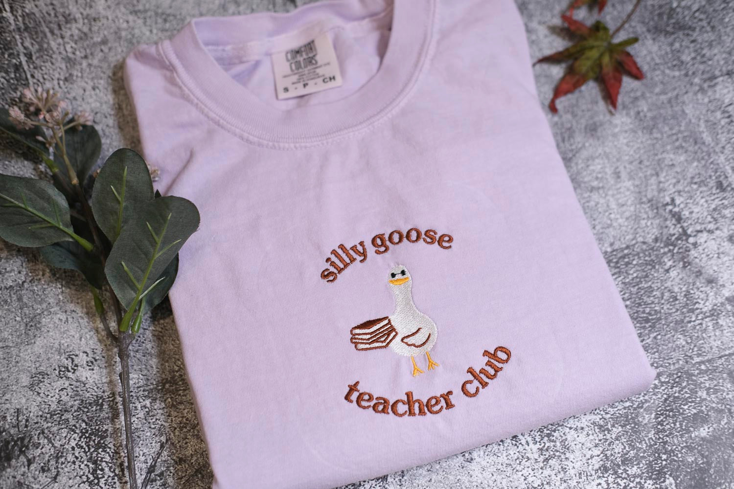 A white t-shirt with a cartoon goose and the text "silly goose teacher club" printed on it, along with a small leaf and a plant.
