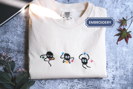 a white t-shirt with three small black creatures with colorful eyes and arms, arranged in a row on the front of the shirt.