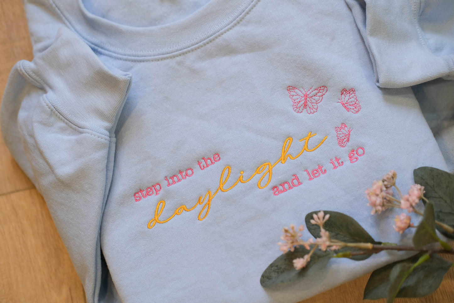 A light blue t-shirt with the text "step into the daylight and let it go" embroidered on it, along with two pink butterflies. The t-shirt is placed on a wooden surface, and there are some pink flowers nearby.