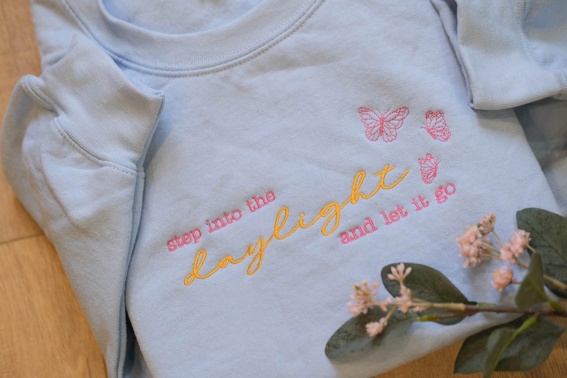 A light blue t-shirt with the text "step into the daylight and let it go" embroidered on it, along with two pink butterflies. The t-shirt is placed on a wooden surface, and there are some pink flowers nearby.