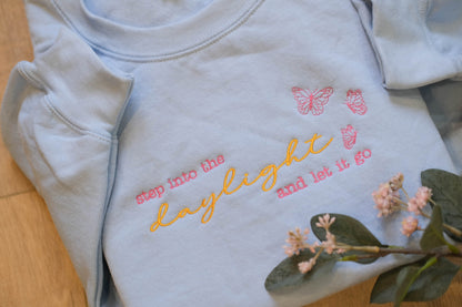 A light blue t-shirt with the text "step into the daylight and let it go" embroidered on it, along with two pink butterflies. The t-shirt is placed on a wooden surface, and there are some pink flowers nearby.