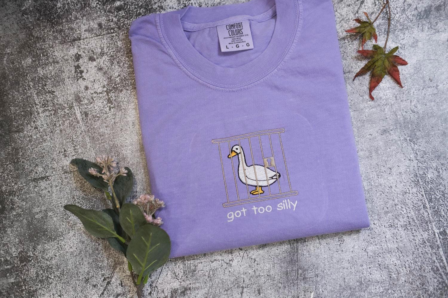 a purple t-shirt with a cartoon duck inside a cage, accompanied by the text &quot;got too silly&quot; printed on it.