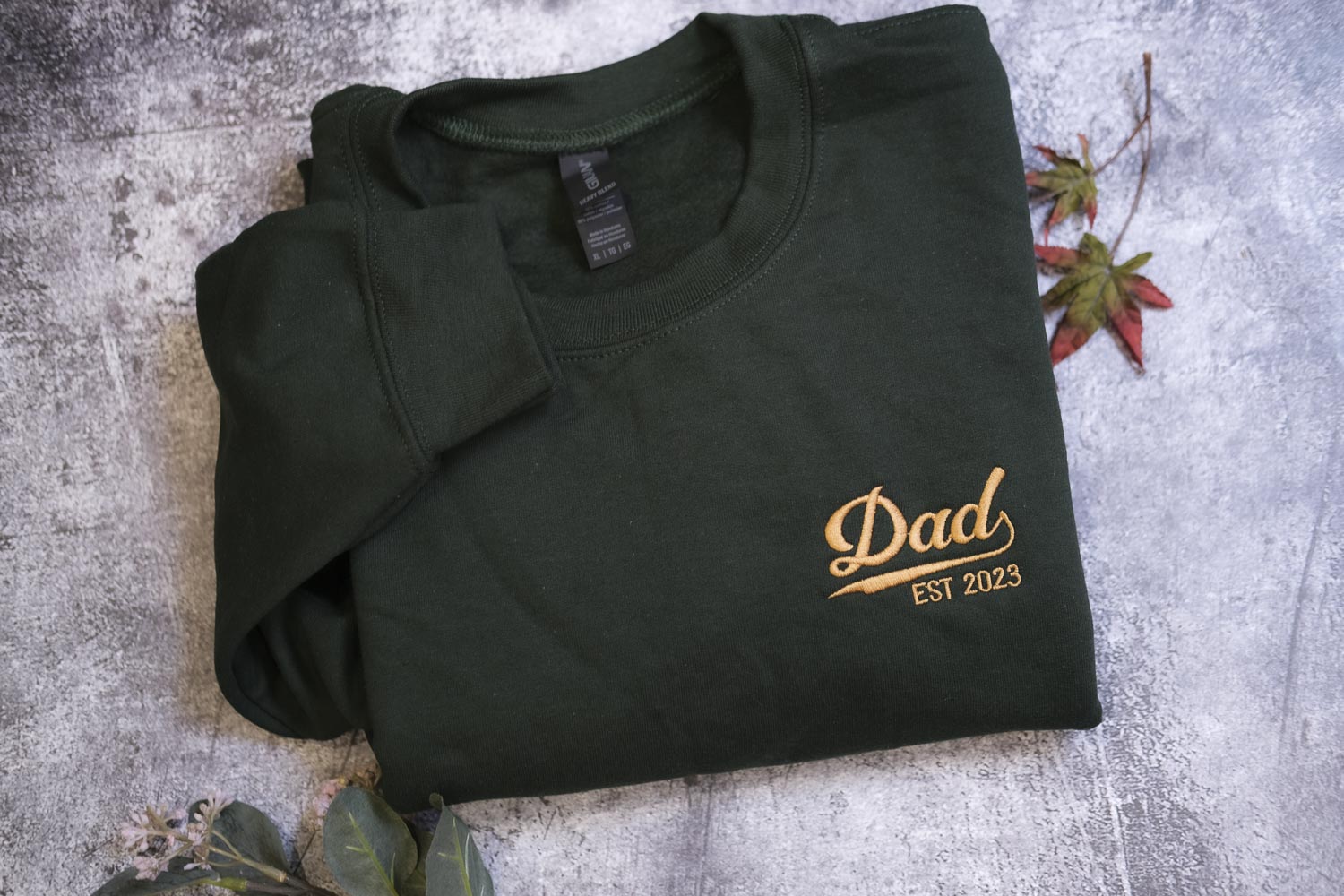 A dark green t-shirt with a gold text logo that says "DAD EST 2013" printed on it, along with some fallen leaves on a gray surface.