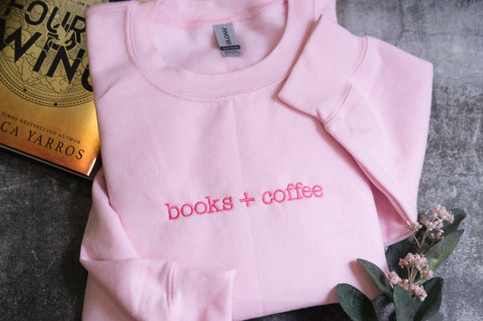 A pink sweatshirt with the text "books + coffee" printed on it, along with a book titled "FOUR WINDS" and a small plant with pink flowers.