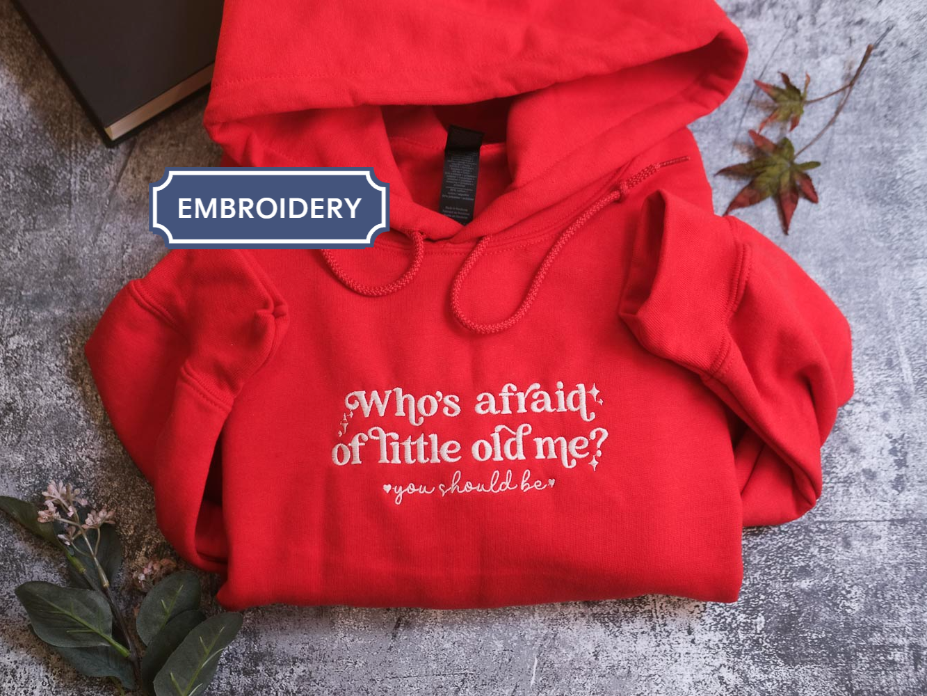 A red hoodie with white text is displayed on a gray surface, accompanied by a black book and some leaves.