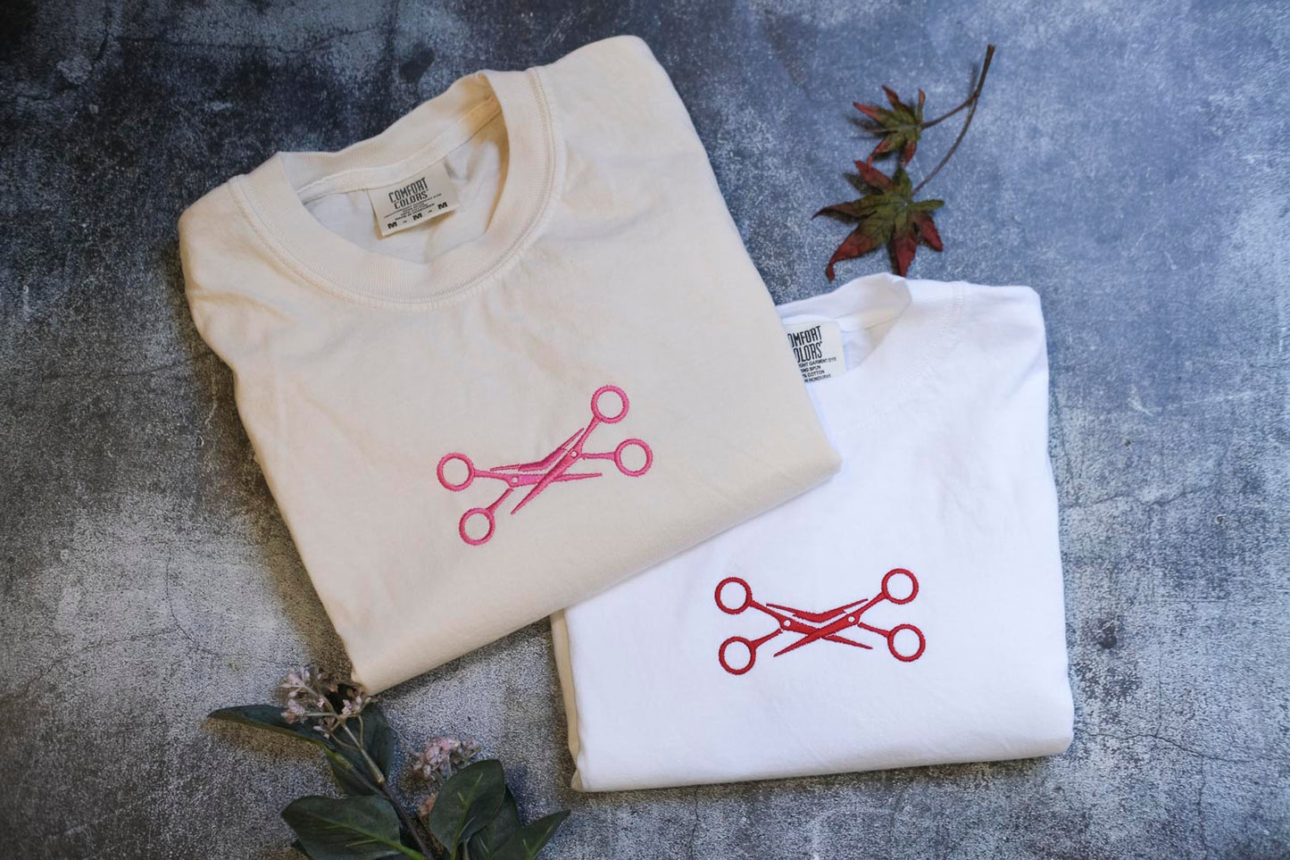 two white t-shirts with red scissors designs, laid out on a gray surface. There are also some leaves and a small plant in the background.
