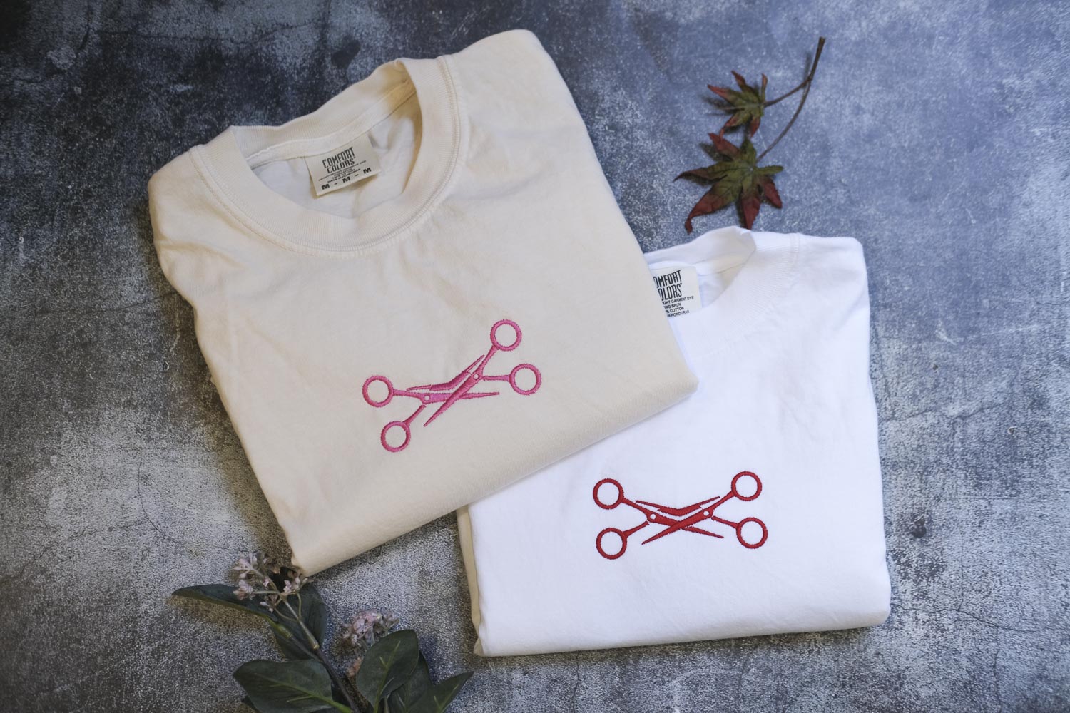 two white t-shirts with red scissors designs, laid out on a gray surface. There are also some leaves and a small plant in the background.