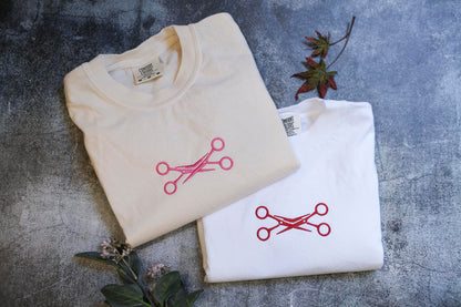 two white t-shirts with red scissors designs, laid out on a gray surface. There are also some leaves and a small plant in the background.