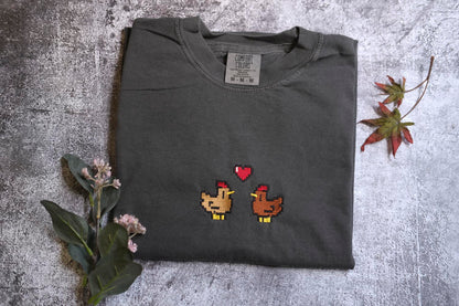 a gray t-shirt with a pixelated design of two chickens, one brown and one yellow, with a heart between them. The t-shirt is placed on a gray surface, and there are some flowers and a leaf nearby.
