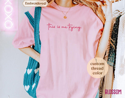 A person wearing a pink t-shirt with the text "This is me trying" printed on it, standing in front of a clothing rack.