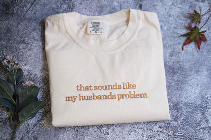 A white t-shirt with the text "that sounds like my husband's problem" printed on it, along with a small label on the collar.