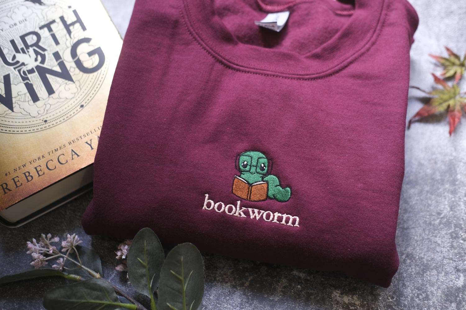 a maroon sweatshirt with a green book worm embroidered on it, along with a book titled "The Earthling" by Rebecca Yay.