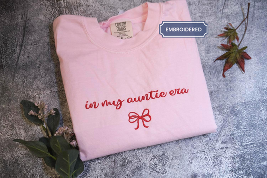 A pink t-shirt with the text &quot;in my auntie era&quot; printed on it, along with a red bow design, is displayed on a gray surface.