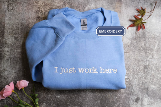 A blue sweatshirt with the text "I just work here" printed on it, along with a small leaf and a pink flower.