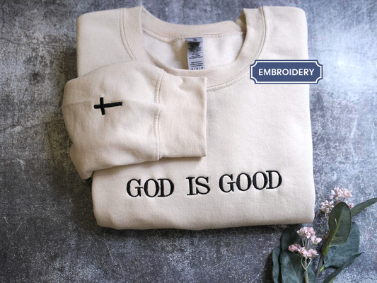 A beige sweatshirt with the text "GOD IS GOOD" printed on it, along with a small black cross symbol, is displayed on a gray surface.