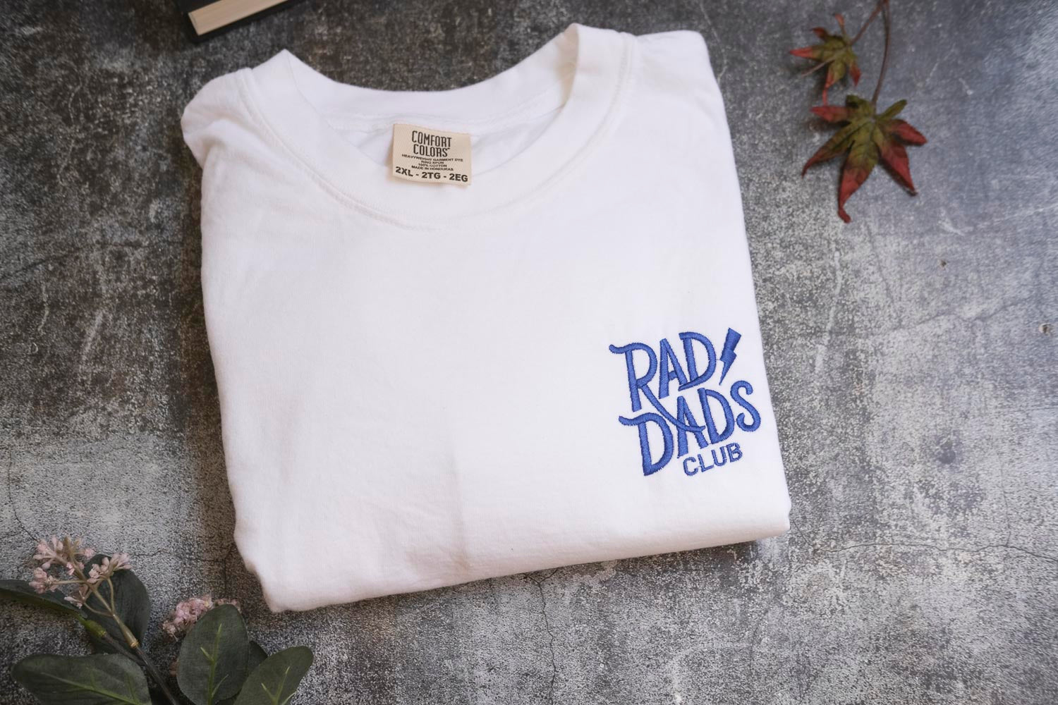 a white t-shirt with blue text and logo, laid out on a gray surface. There are some leaves and a small plant in the background.