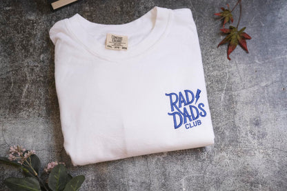 a white t-shirt with blue text and logo, laid out on a gray surface. There are some leaves and a small plant in the background.