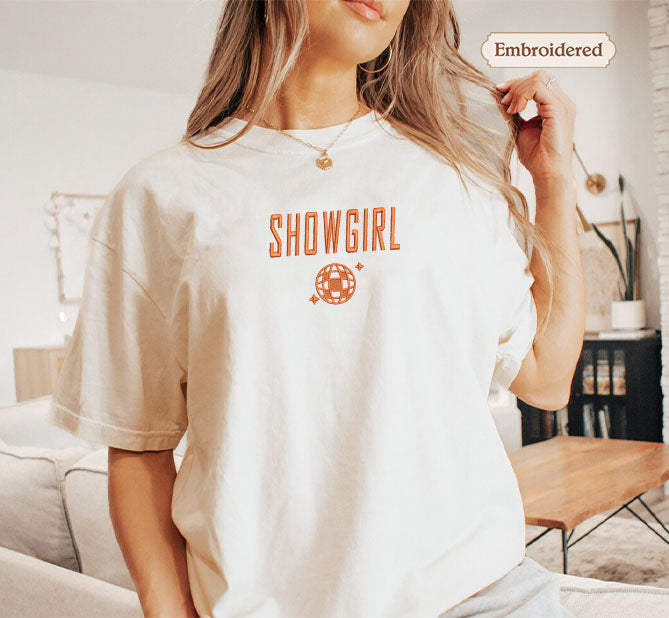 A woman wearing a white t-shirt with the text "SHOWGIRL" printed on it, posing in a room with a couch and a coffee table.