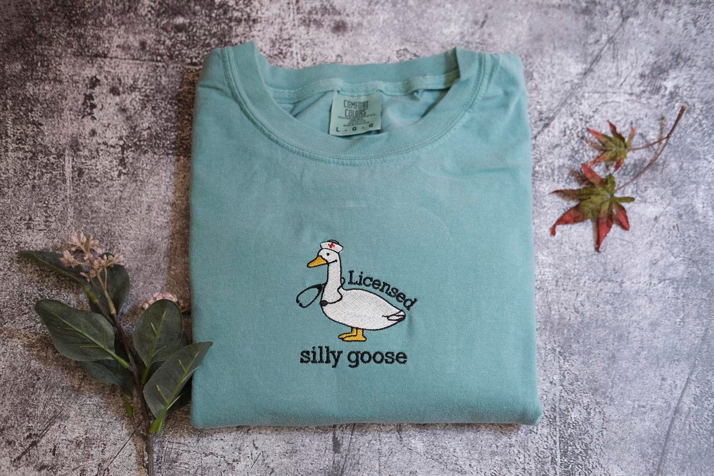 A light blue t-shirt with a cartoon image of a duck wearing a nurse's outfit and the text "Licensed silly goose" printed on it, along with some leaves and a small flower.