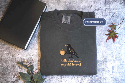 a black t-shirt with a graphic design of a bird holding a cup, accompanied by the text &quot;Hello darkness, my old friend&quot; printed on it. The t-shirt is placed on a gray surface, with a black book and a plant nearby.