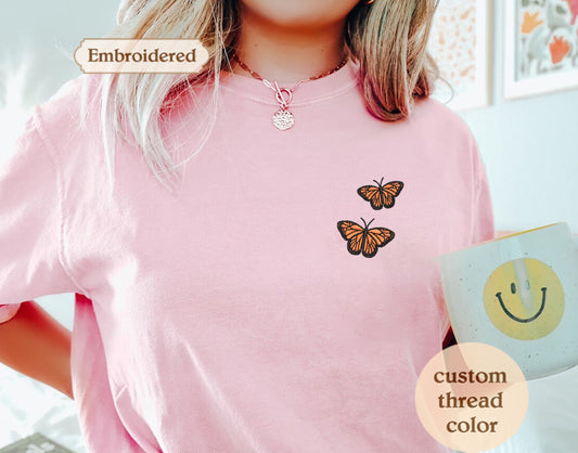 A person wearing a pink t-shirt with a butterfly embroidered on it, holding a mug with a smiley face design.