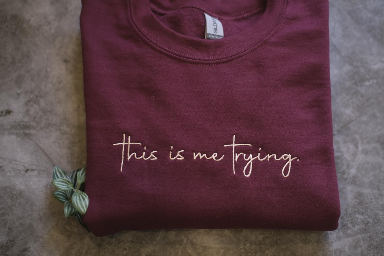 A maroon t-shirt with the text "This is me trying" printed on it, along with a small green plant or leaf.