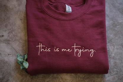 A maroon t-shirt with the text "This is me trying" printed on it, along with a small green plant or leaf.