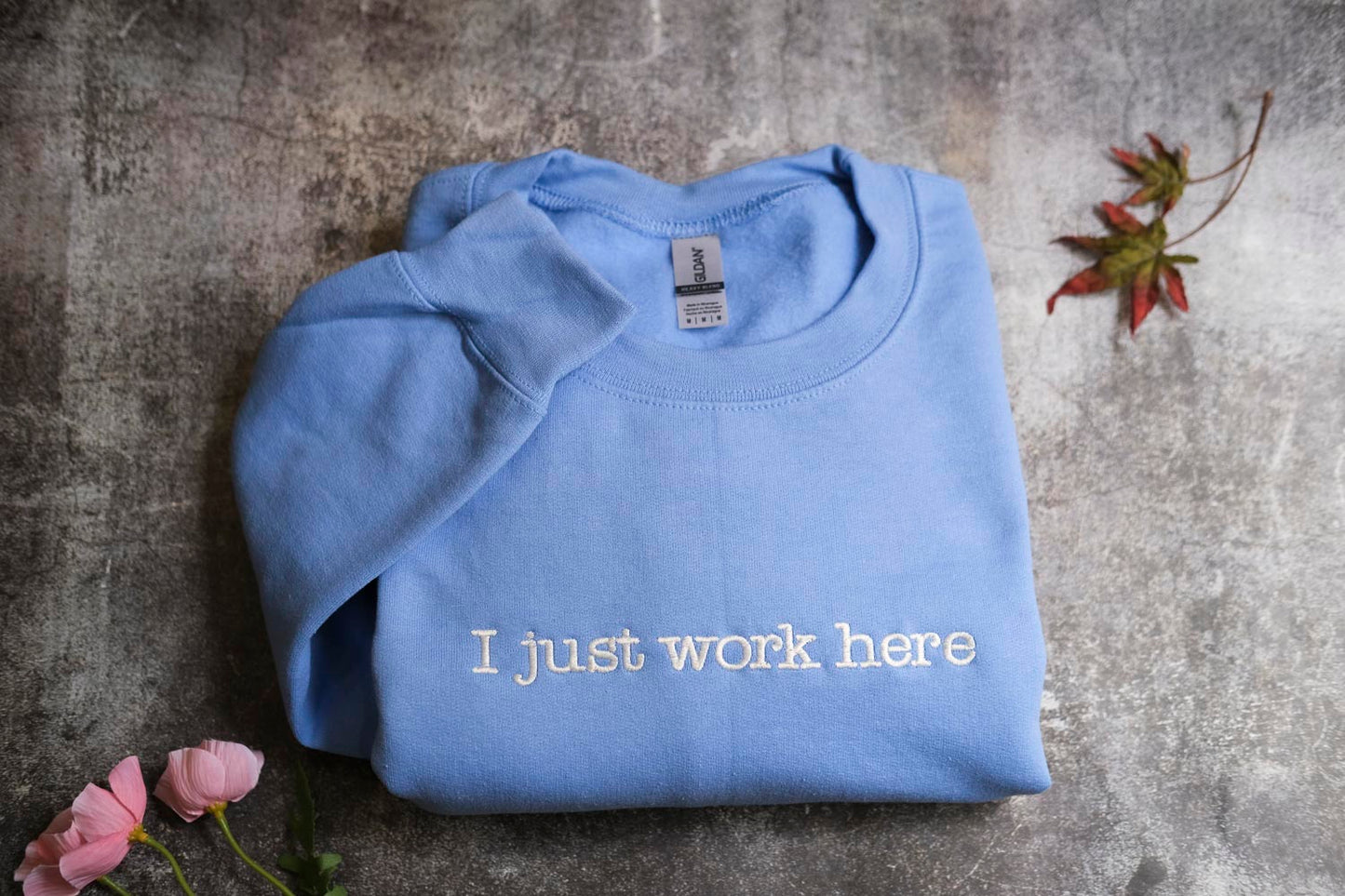 A blue t-shirt with the text "I just work here" printed on it, along with some pink flowers and a fallen leaf on a textured gray surface.