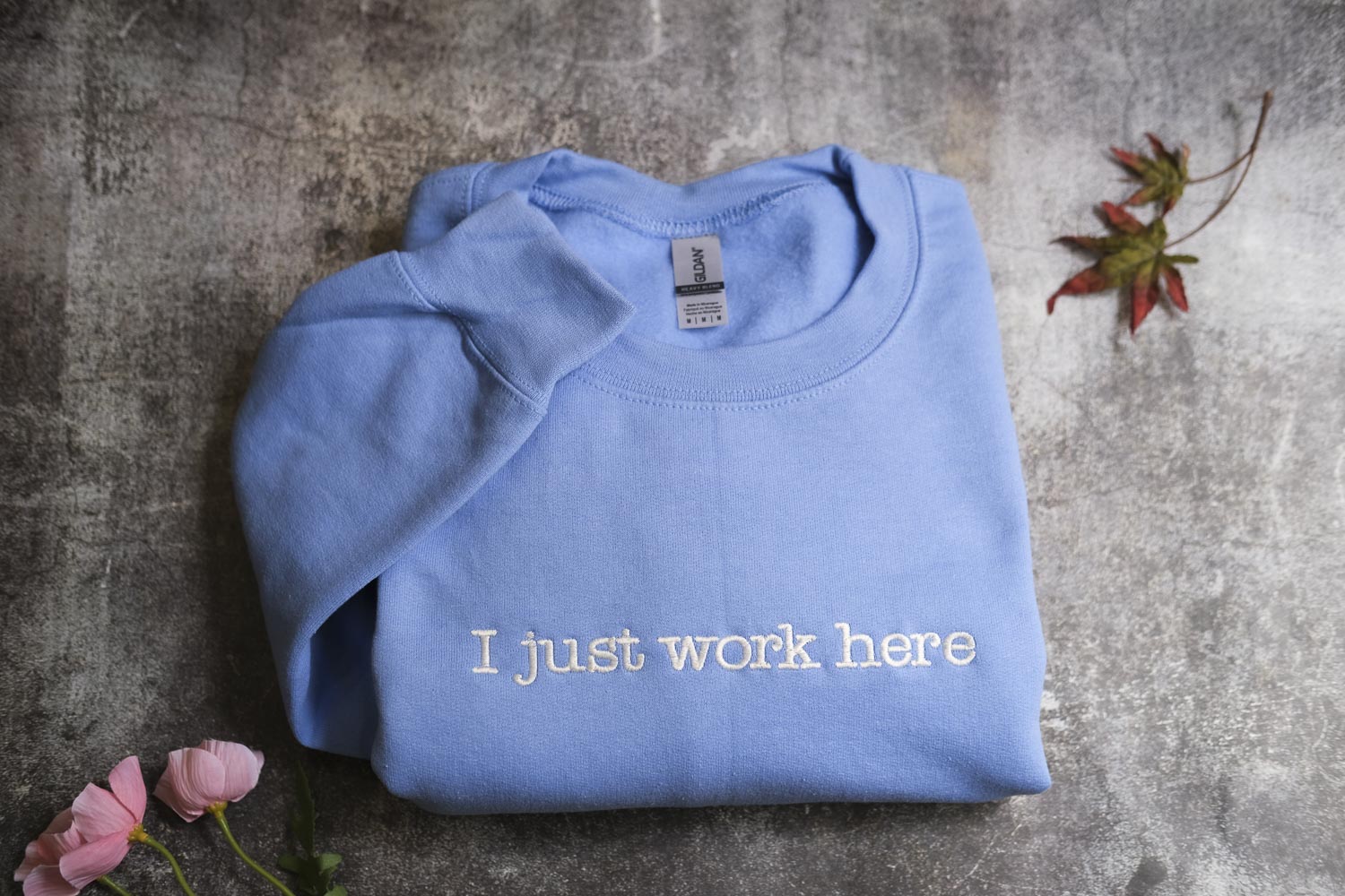 A blue t-shirt with the text "I just work here" printed on it, along with some pink flowers and a fallen leaf on a textured gray surface.