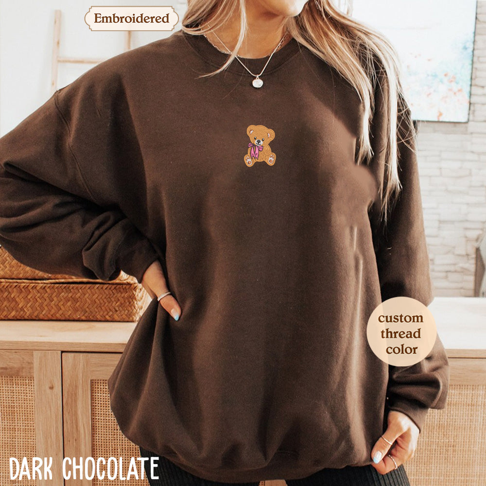 A woman wearing a brown sweater with a teddy bear embroidered on it, standing in front of a wooden cabinet.