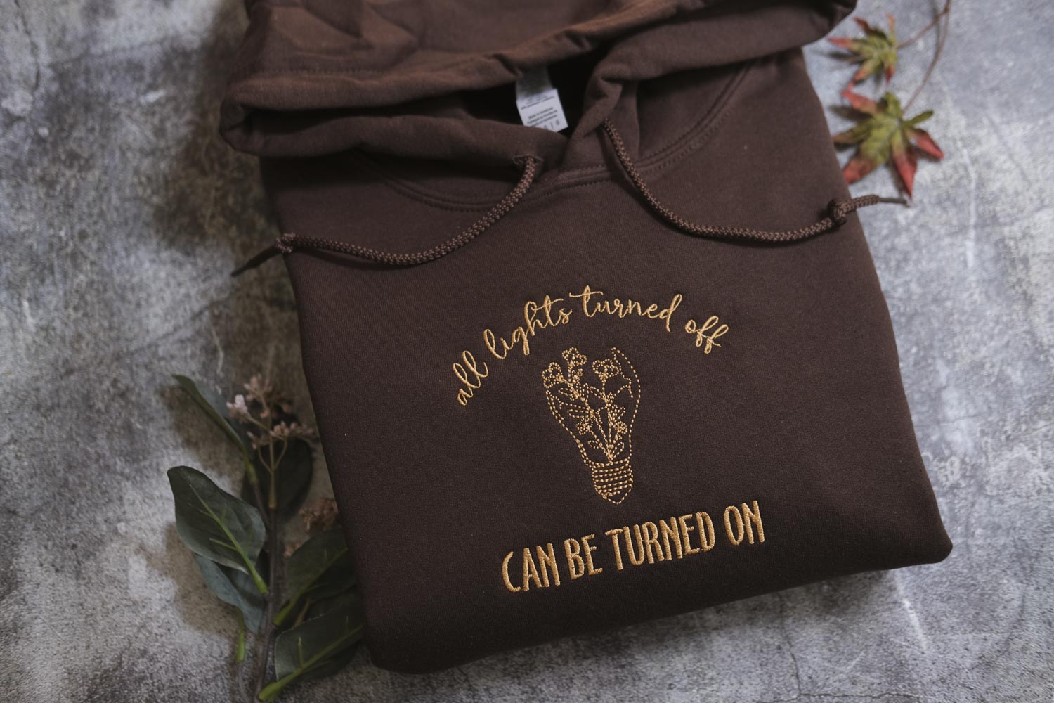A brown hoodie with a gold embroidered design and text on it, placed on a gray surface with some leaves and a plant.