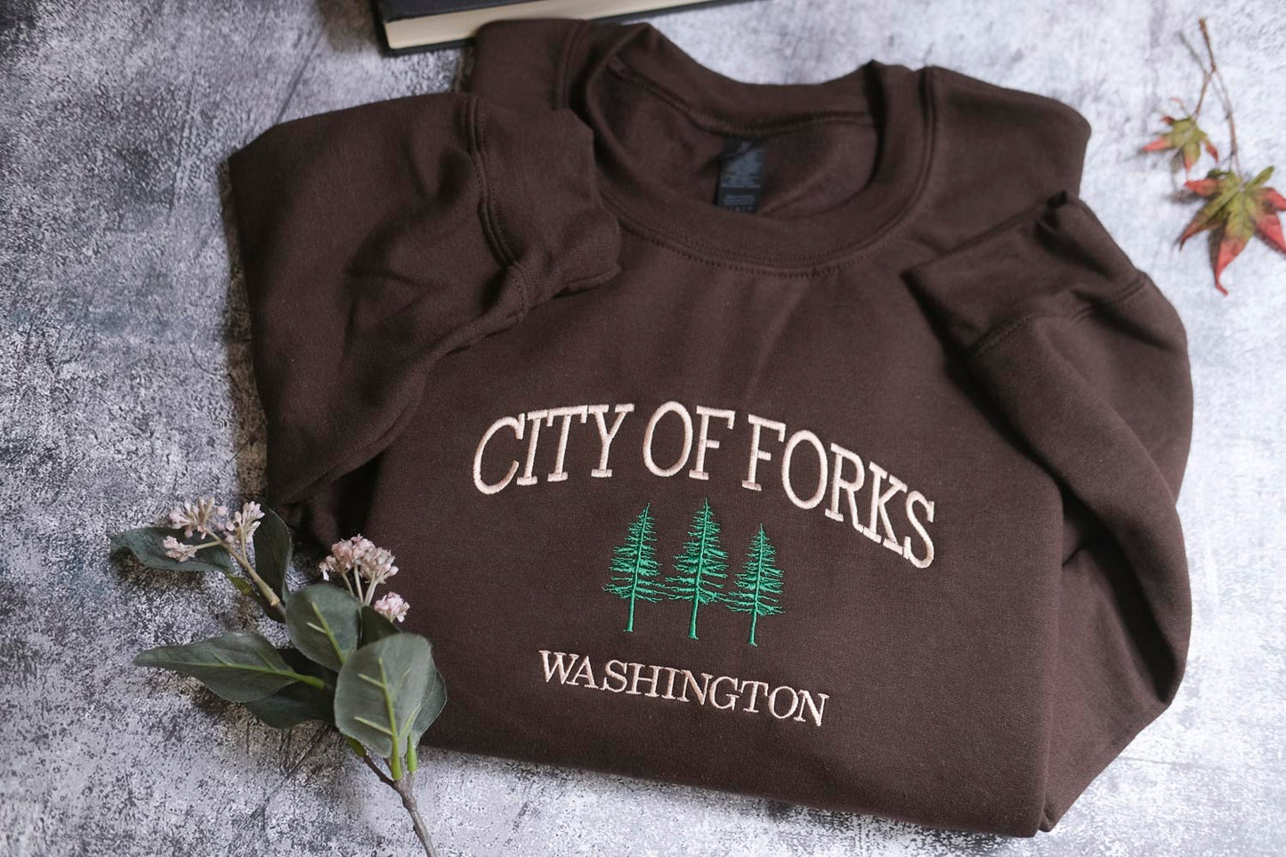 A brown sweatshirt with the text "CITY OF FORKS WASHINGTON" printed on it, along with a graphic of three pine trees. The sweatshirt is laid out on a gray surface, and there are some small white flowers and a leaf nearby.