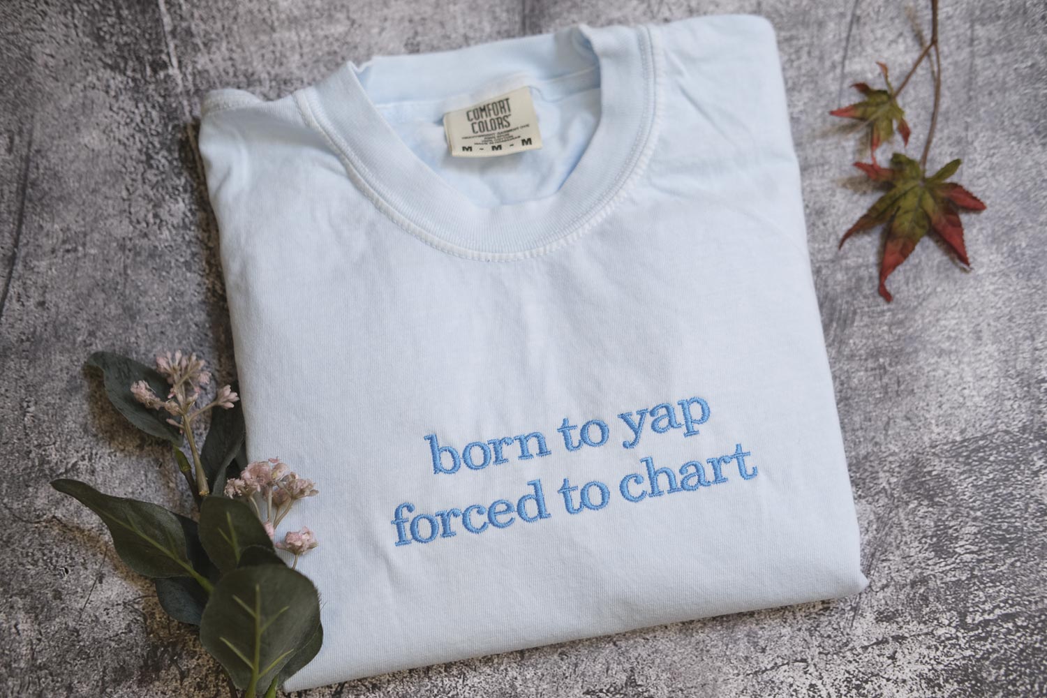 A white t-shirt with the text "born to yap forced to chart" printed on it, along with some leaves and flowers.