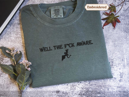 a gray t-shirt with the text "WELL THE F*CK AWARE" printed on it, along with a graphic of a dragon. The t-shirt is placed on a surface with some leaves and flowers, and there is a small embroidered label visible on the collar of the shirt.