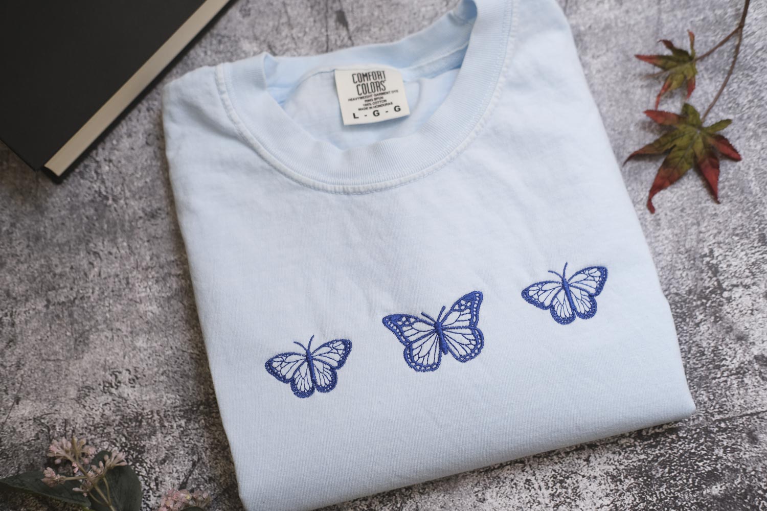 a white t-shirt with three blue butterflies printed on it, placed on a gray surface alongside some leaves and a book.