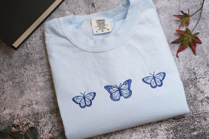 a white t-shirt with three blue butterflies printed on it, placed on a gray surface alongside some leaves and a book.