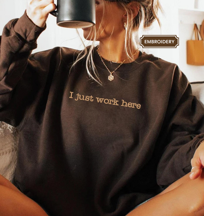 A woman wearing a brown sweatshirt with the text "I just work here" embroidered on it, holding a coffee mug and sitting on a couch.