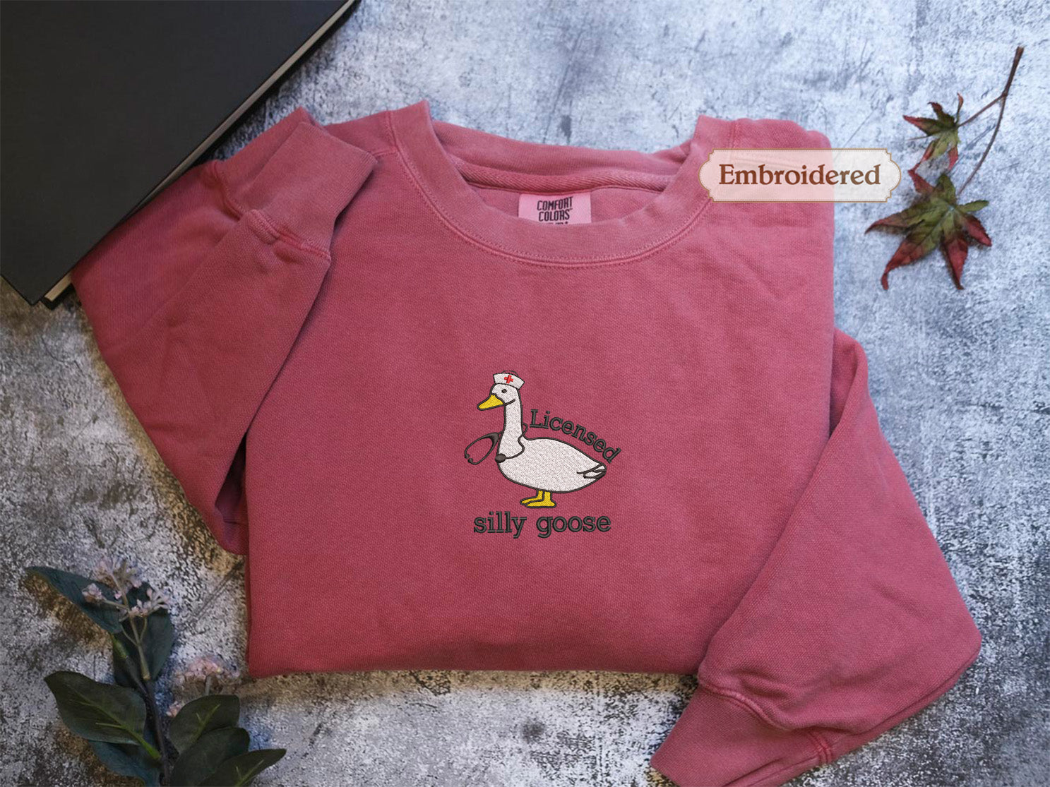 a red long-sleeved shirt with a cartoon duck and the text "Licenced silly goose" embroidered on it. The shirt is placed on a gray surface, with a leaf and a book visible in the background.