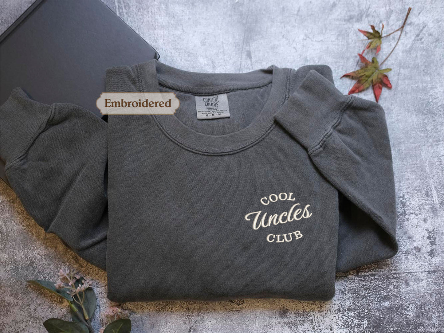 a gray long-sleeved shirt with the text "Cool Uncle Club" embroidered on it, along with a label that reads "Embroidered". The shirt is placed on a gray surface, and there are some leaves and a book visible in the background.
